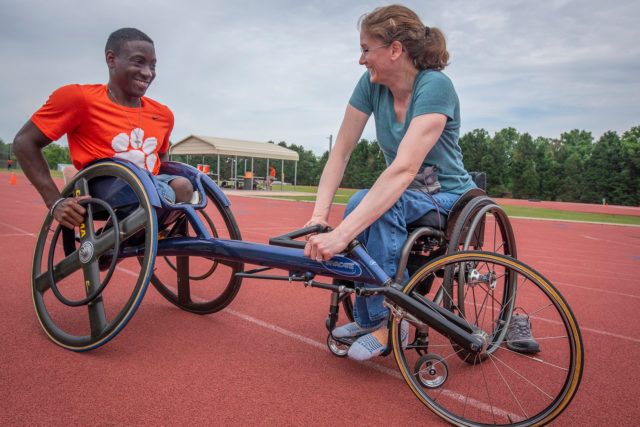 Two people in wheelchairs sit on either side of a racing wheelchair, with a cloudy sky behind them.