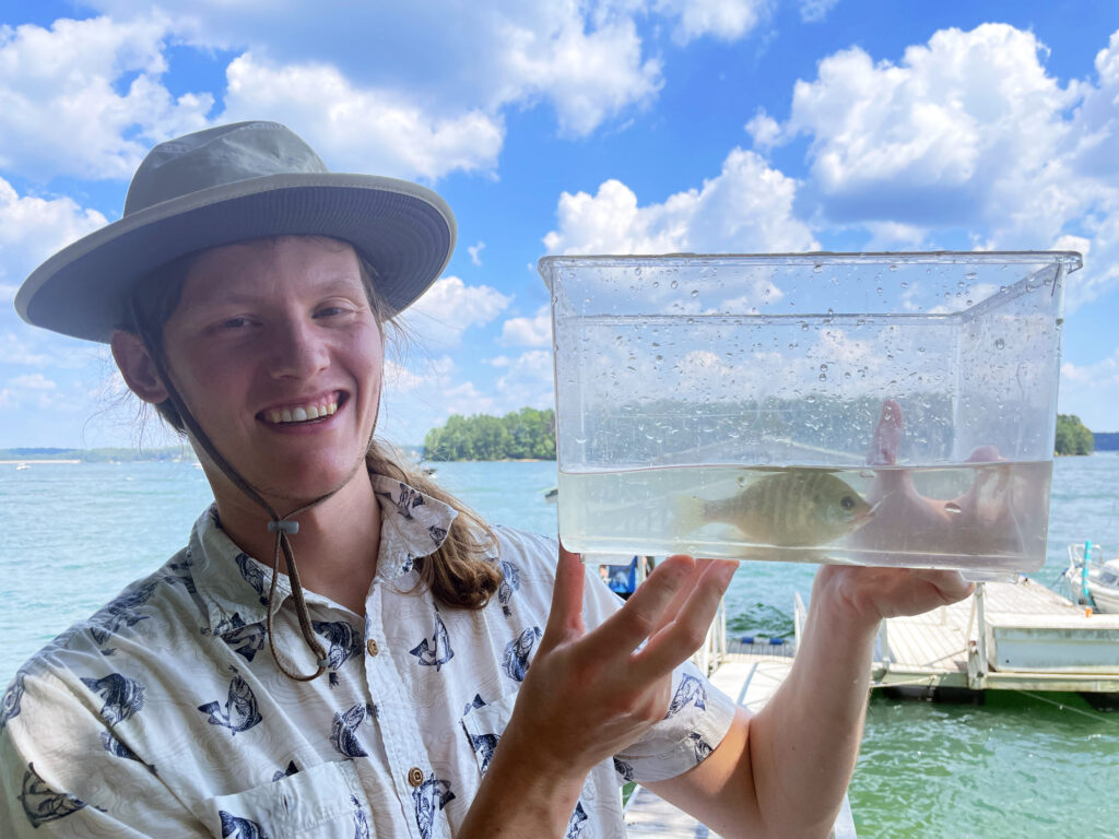 Man standing on dock holding a clear container containing a fish.