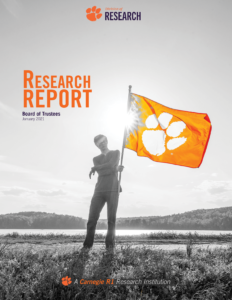 The photo shows a Clemson student holding an orange Clemson flag with a white tiger paw. The image includes the text "Research Report, Board of Trustees, January 2021". Click the photo to view the report. 