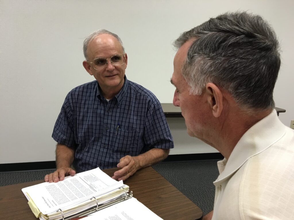 Two men are sitting at a table, looking at a binder of materials.