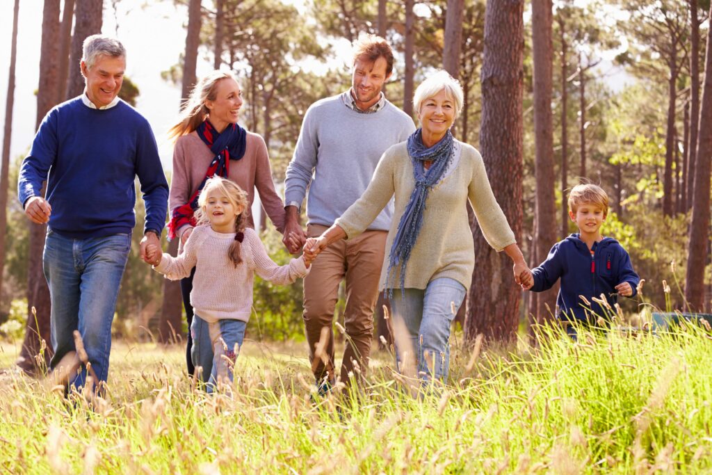A multi-generational family walking hand-in-hand through a sunlit forest clearing. The group includes two older adults, two middle-aged adults, and two young children, all dressed in casual fall clothing with trees and tall grass surrounding them.
