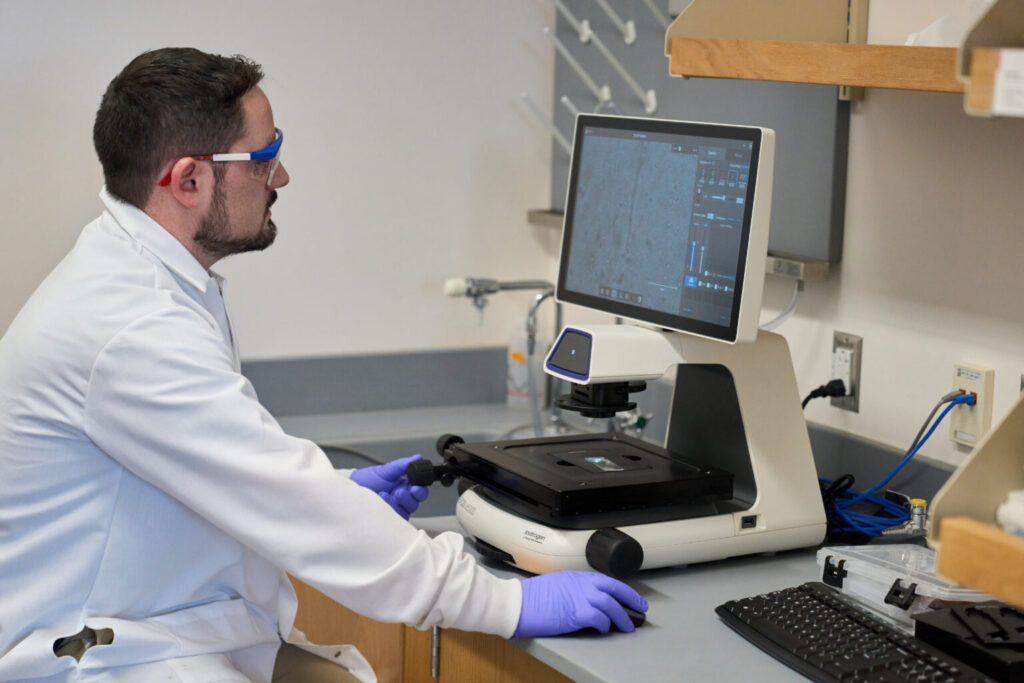 scientist with goggles on looking into a microscopic computer screen with latex gloves on.