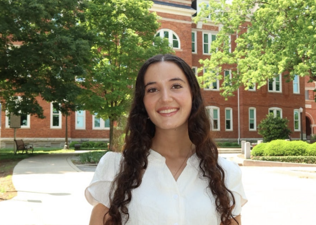 headshot of girl with brunette curly long hair in front of red brick building.