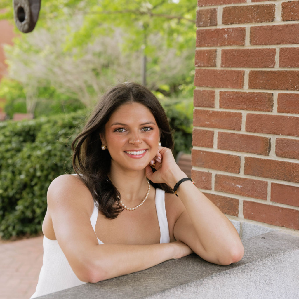 Woman in white dress with brown hair leaning on stone with arms. 