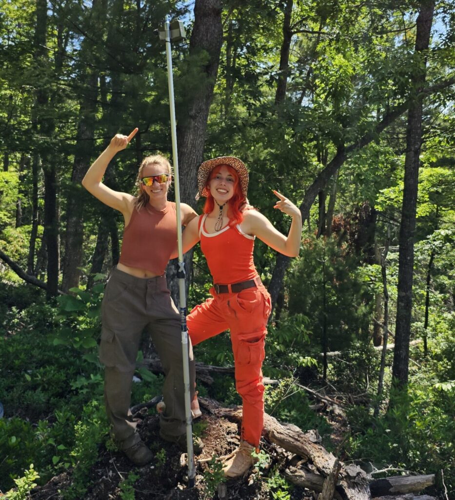 Two students pointing towards an acoustic recording device on a pole in the forest.