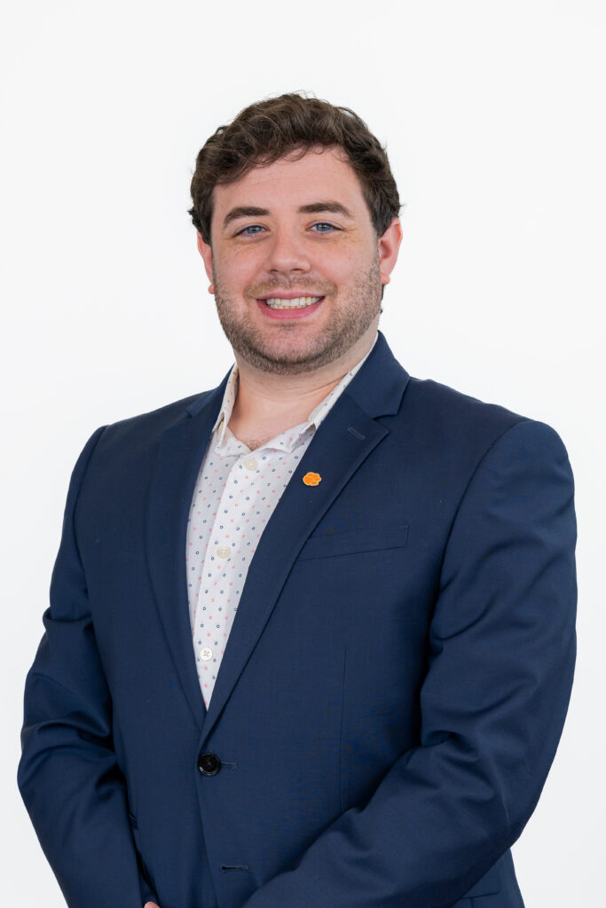 White man in blue suite standing in front of a white background smiling