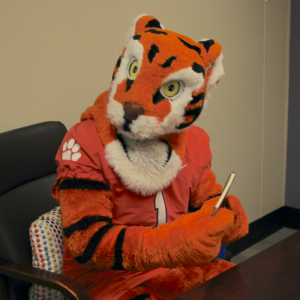 The Clemson Tiger Mascot sitting at a desk, holding a notebook.