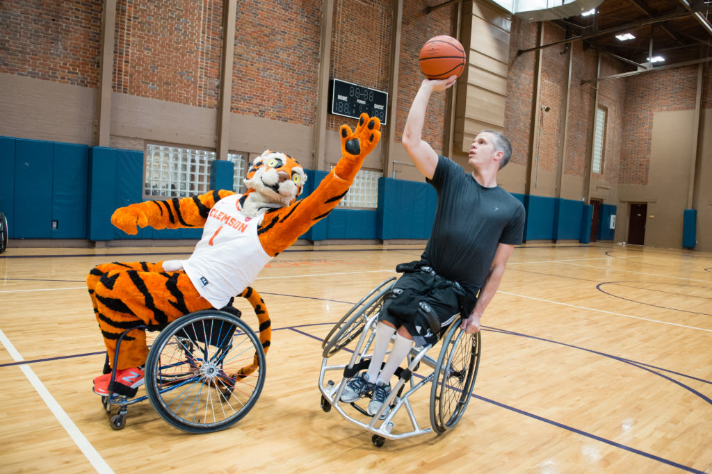 The Clemson Tiger mascot plays basketball, using a wheelchair. He reaches up for a ball held by another player using a wheelchair.