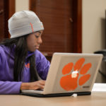 Student in Vickery Hall typing on a mac decorated with a Clemson paw sticker