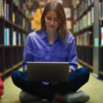 Image depicts a woman sitting on the floor of a library typing on a laptop
