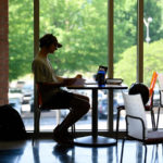Student seated at a table in front of a window working on a laptop
