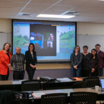 Seven members of the first QM cohort stand in front of a projector showing 3 other participants who joined via Zoom
