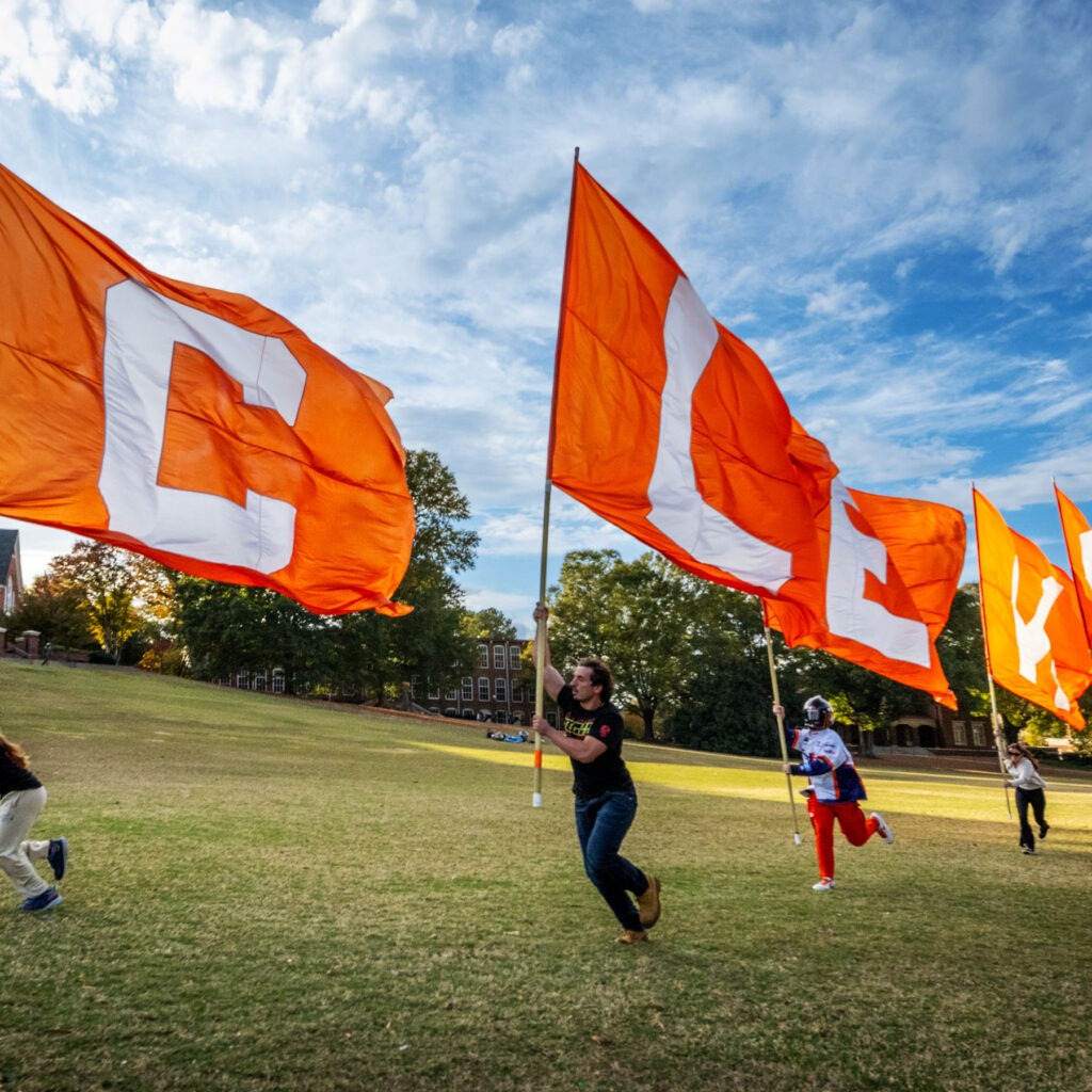 Image of Clemson students running across a field with big orange flags that spell out the word C-L-E-M-S-O-N