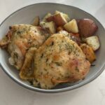 A bowl containing two herb-baked chicken thighs served with roasted potatoes