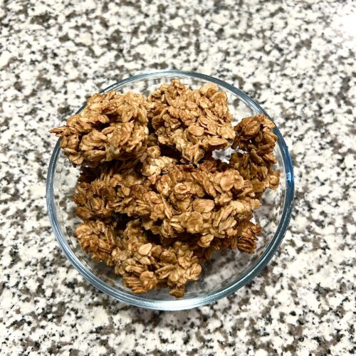 A glass bowl filled with homemade peanut butter granola containing oatmeal, peanut butter, maple syrup, butter, salt, and vanilla.